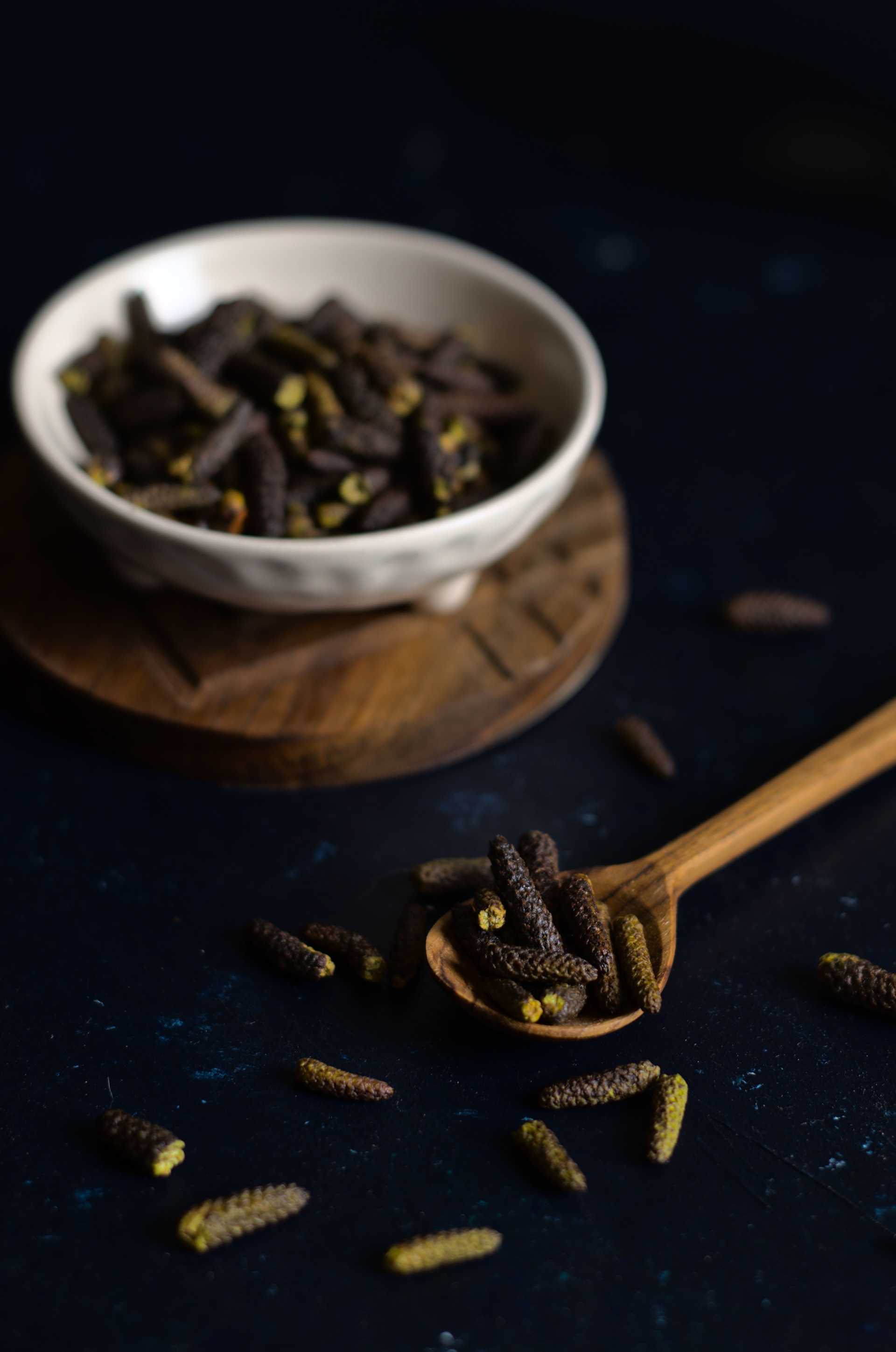 Poivre des dunes fumé dans une petite cuiller de bois sur fond bleu foncé. Derrière, un petit bol blanc contient davantage de poivre des dunes fumé sur un socle de bois.