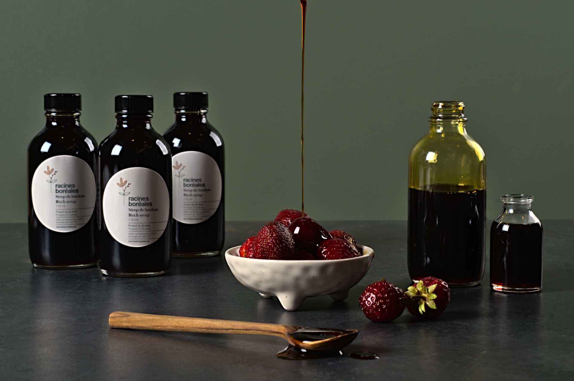 Bouteilles de sirop de bouleau, fraises dans un plat blanc et cuiller de sirop de bouleau sur un comptoir gris-bleu devant un fond vert.