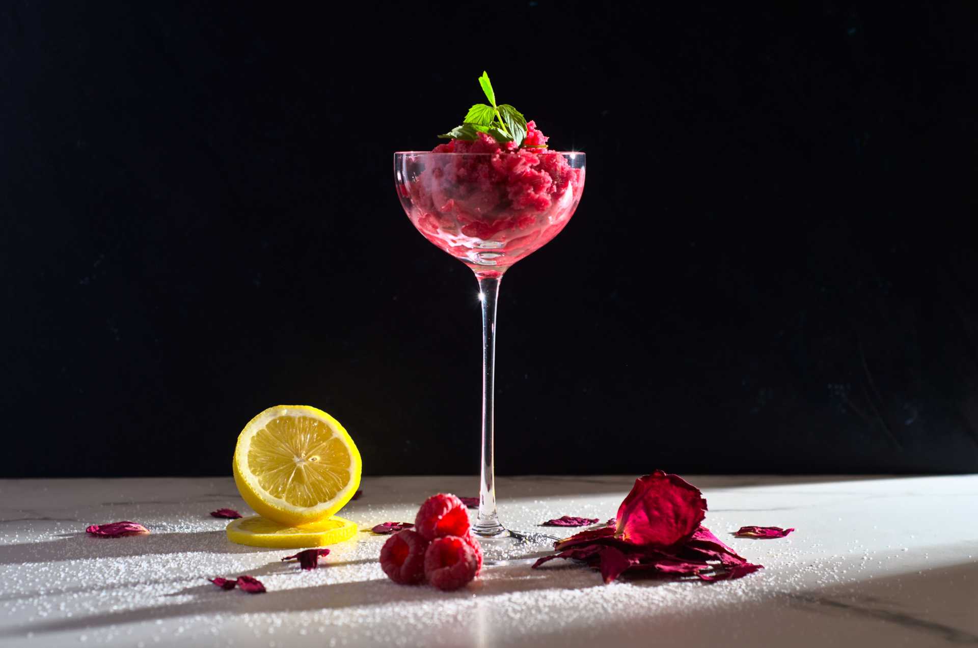 Granité à la framboise et à la rose sauvage (pétales d'églantier) déposé dans une coupe avec une lumière tranchante. Sur le comptoir de marbre sont déposés un citron, des framboises et des pétales de rose sauvage.