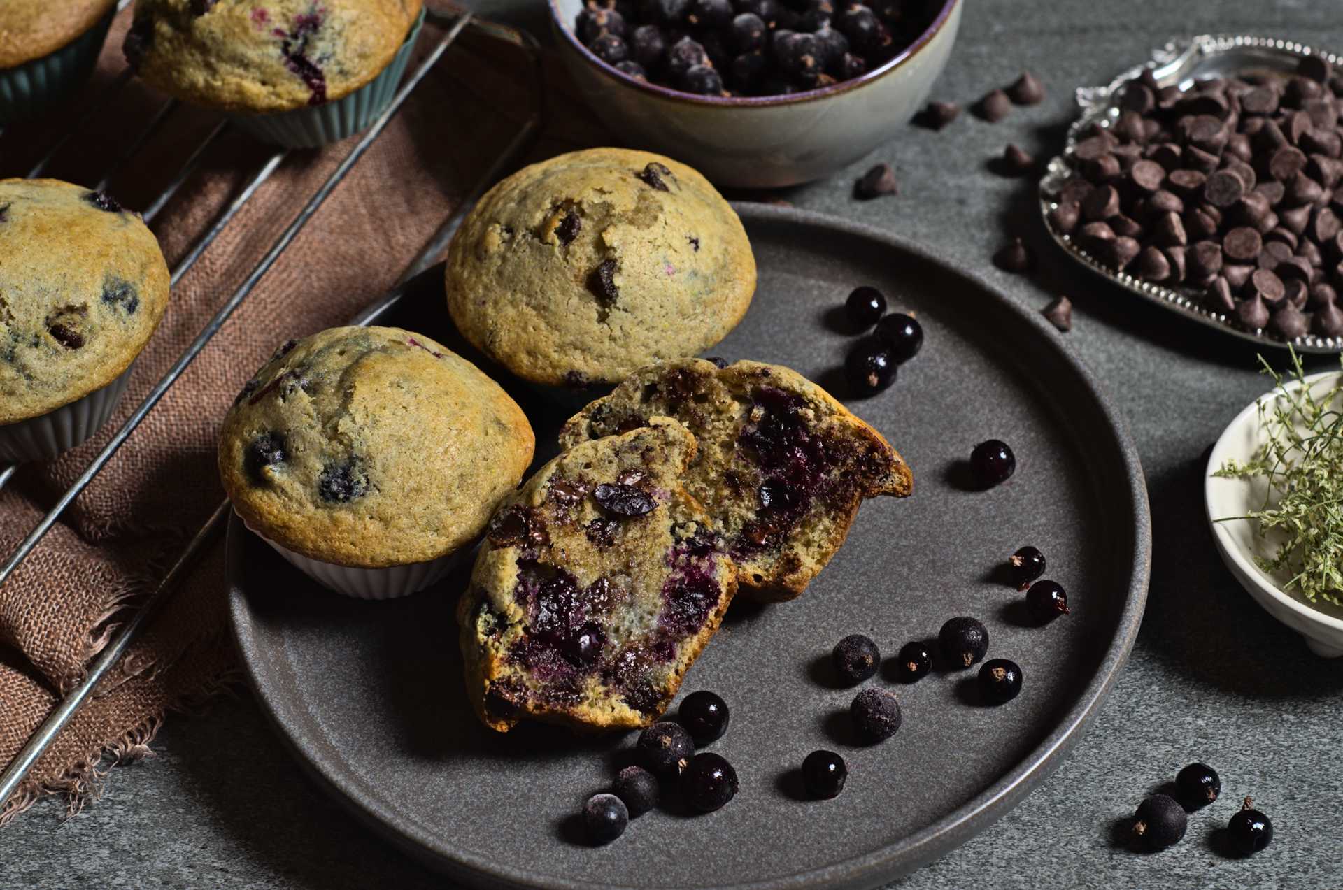 Muffins aux cassis, chocolat et mélilot faits à partir de cassis du Québec congelés de Racines boréales. Les muffins sont disposés dans une assiette grise et entourés d'un bol de cassis, de pépites de chocolat et de fleurs de mélilot.