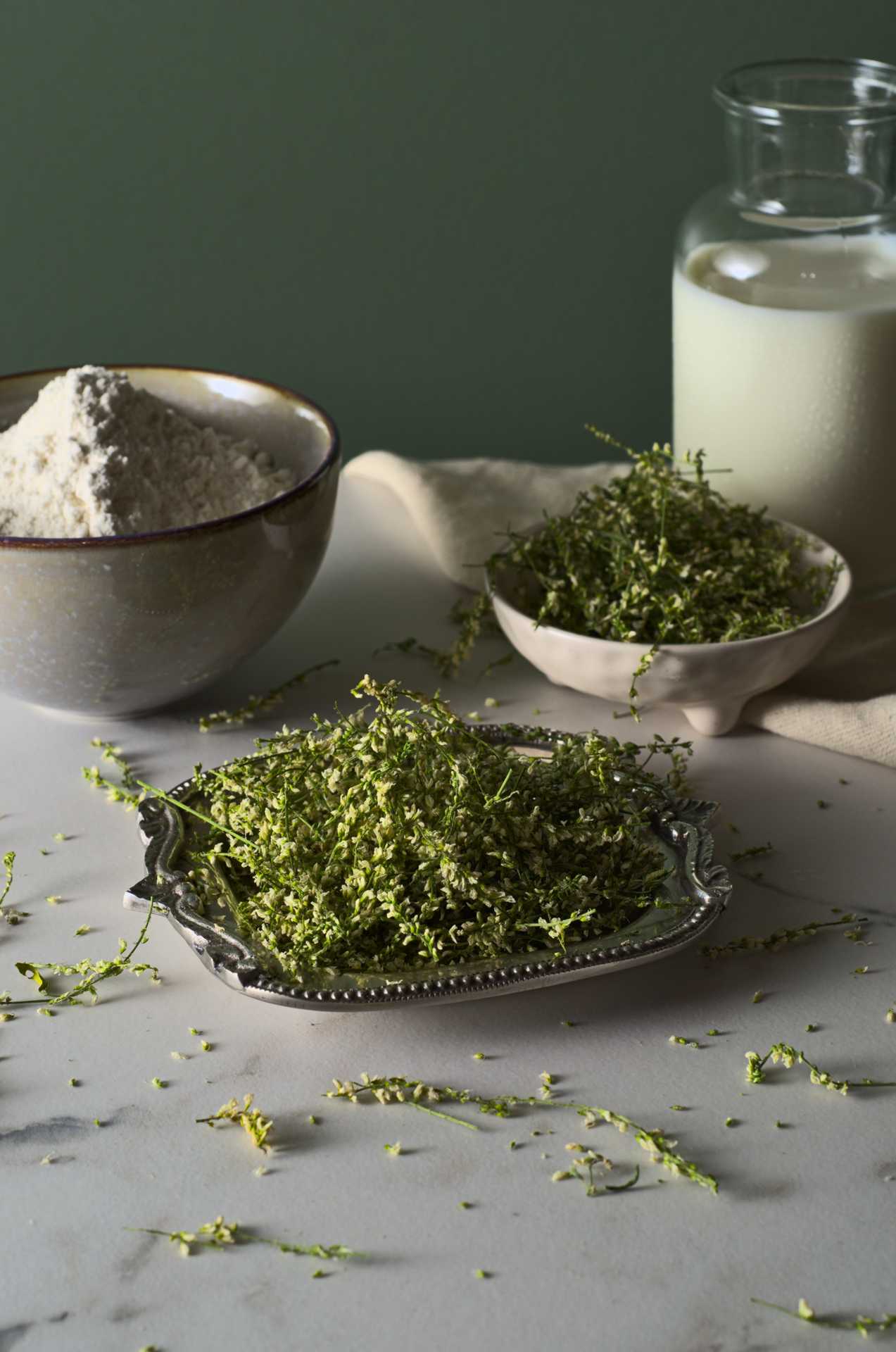 Fleurs et tiges de mélilot séché sur une surface de marbre devant un fond vert. Le melilot est présenté dans un plat métallique accompagné d'un bol de farine, d'une bouteille de lait et d'un autre petit bol rempli de mélilot. Le fond de la photo est vert.