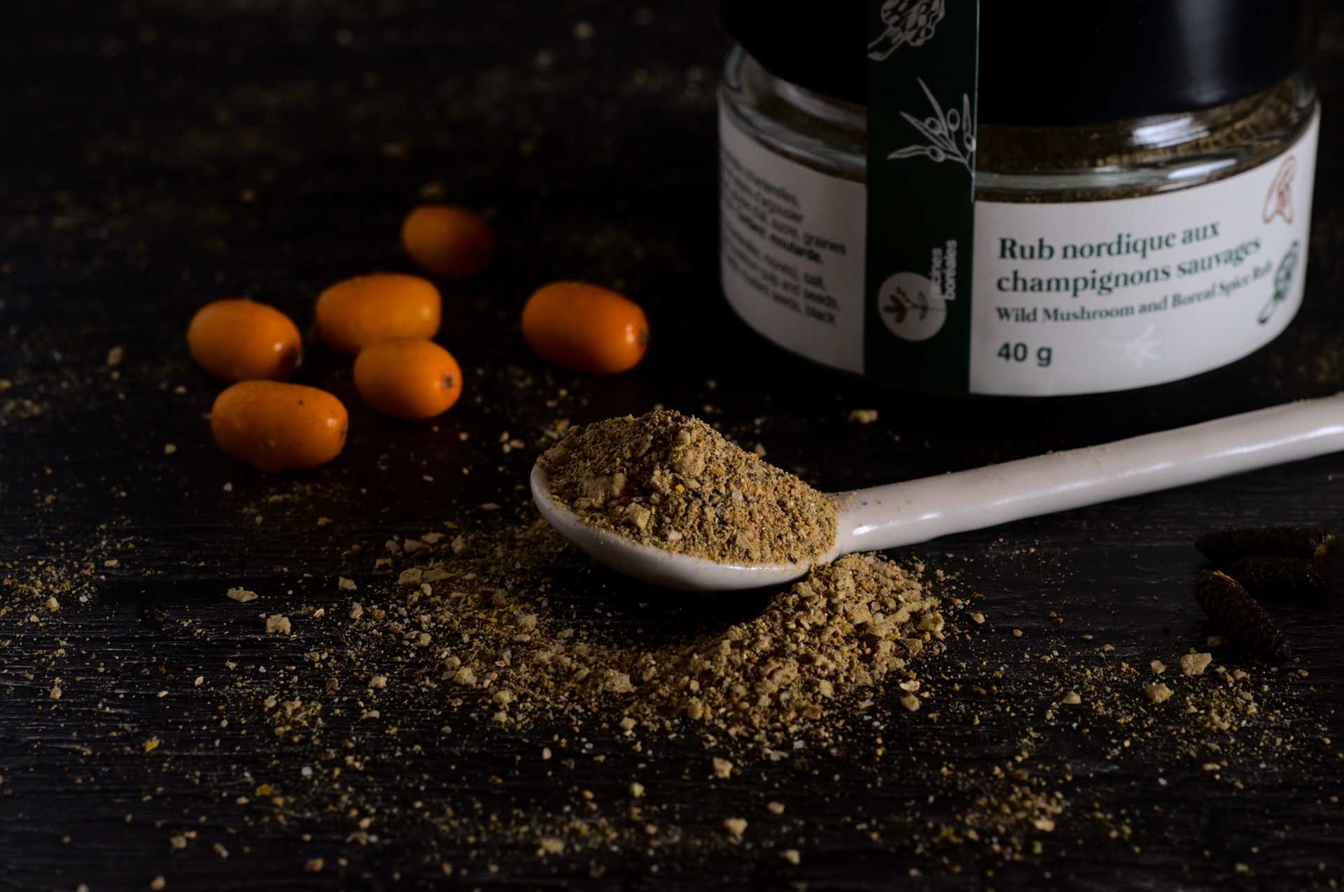 Rub nordique aux champignons sauvages dans une cuillère en céramique blanche sur fond noir. Des baies d'argousier sont disposées près de la cuillère, ainsi que le pot.